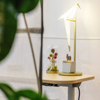A Clérachic bird lamp on a gold stand sits on a pale wooden desk, beside a glass dome of dried flowers and a pen pot.