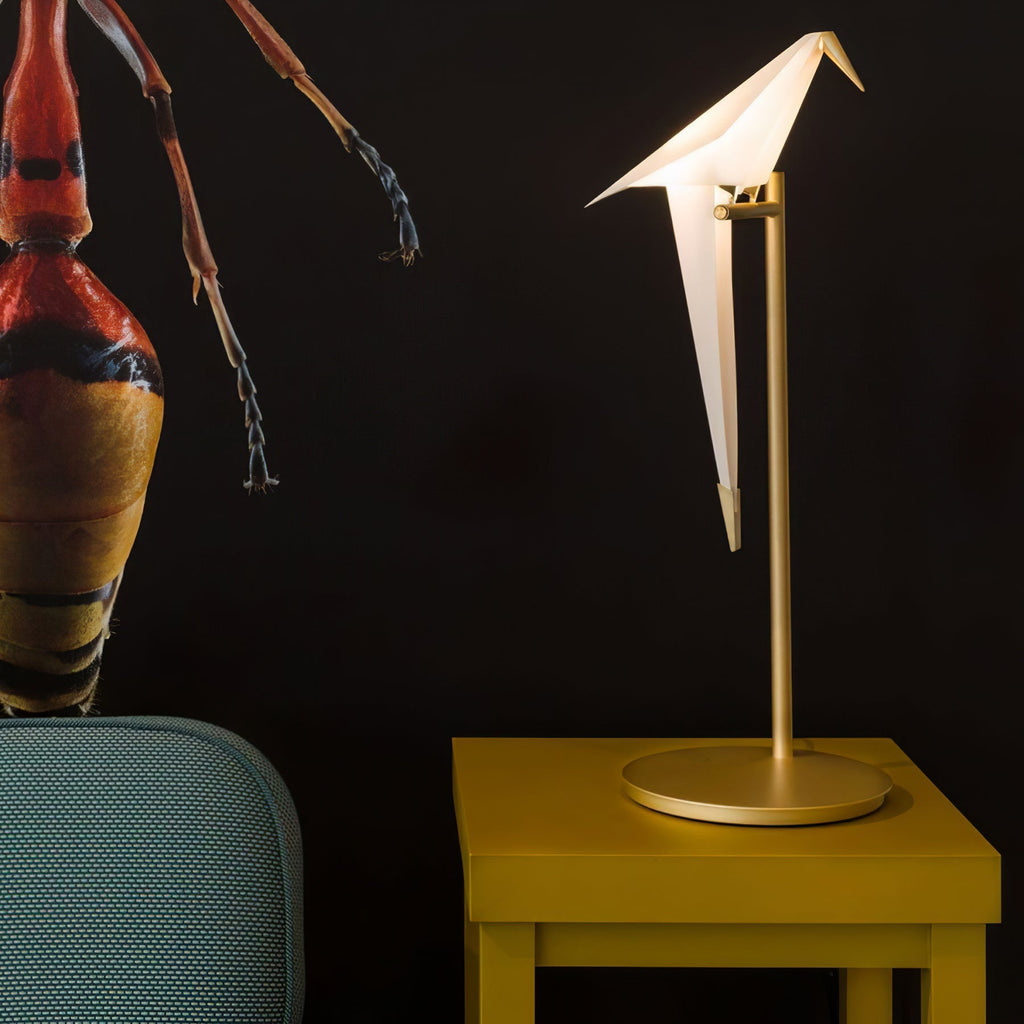 This Clérachic bird table lamp glows on a mustard-yellow side table in front of a dark wall with a red insect artwork.