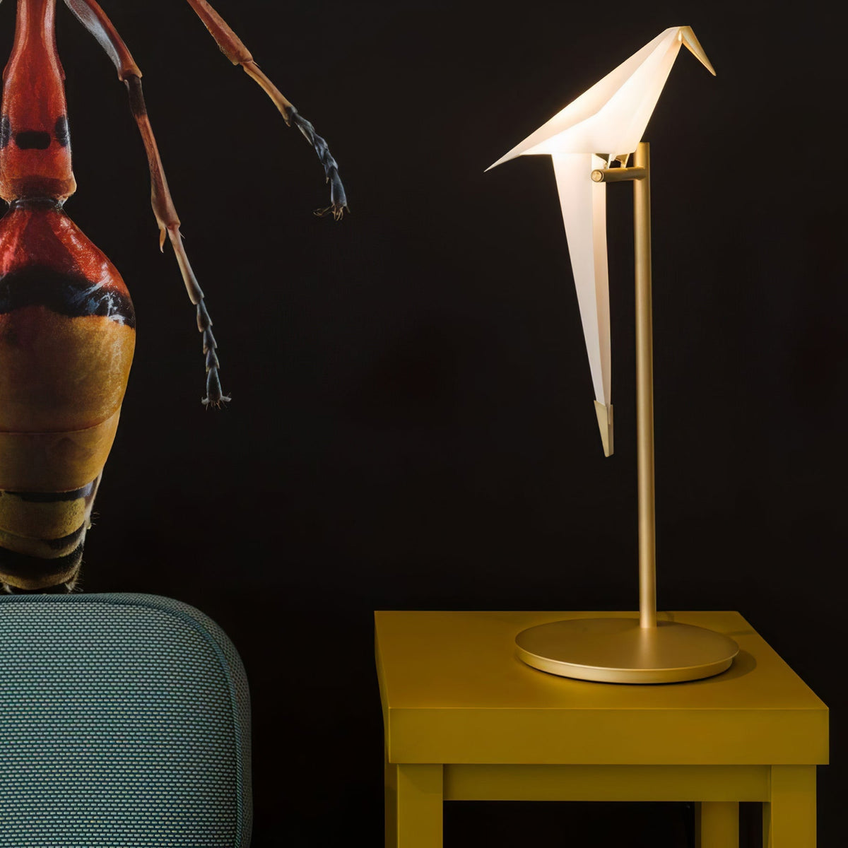This Clérachic bird table lamp glows on a mustard-yellow side table in front of a dark wall with a red insect artwork.