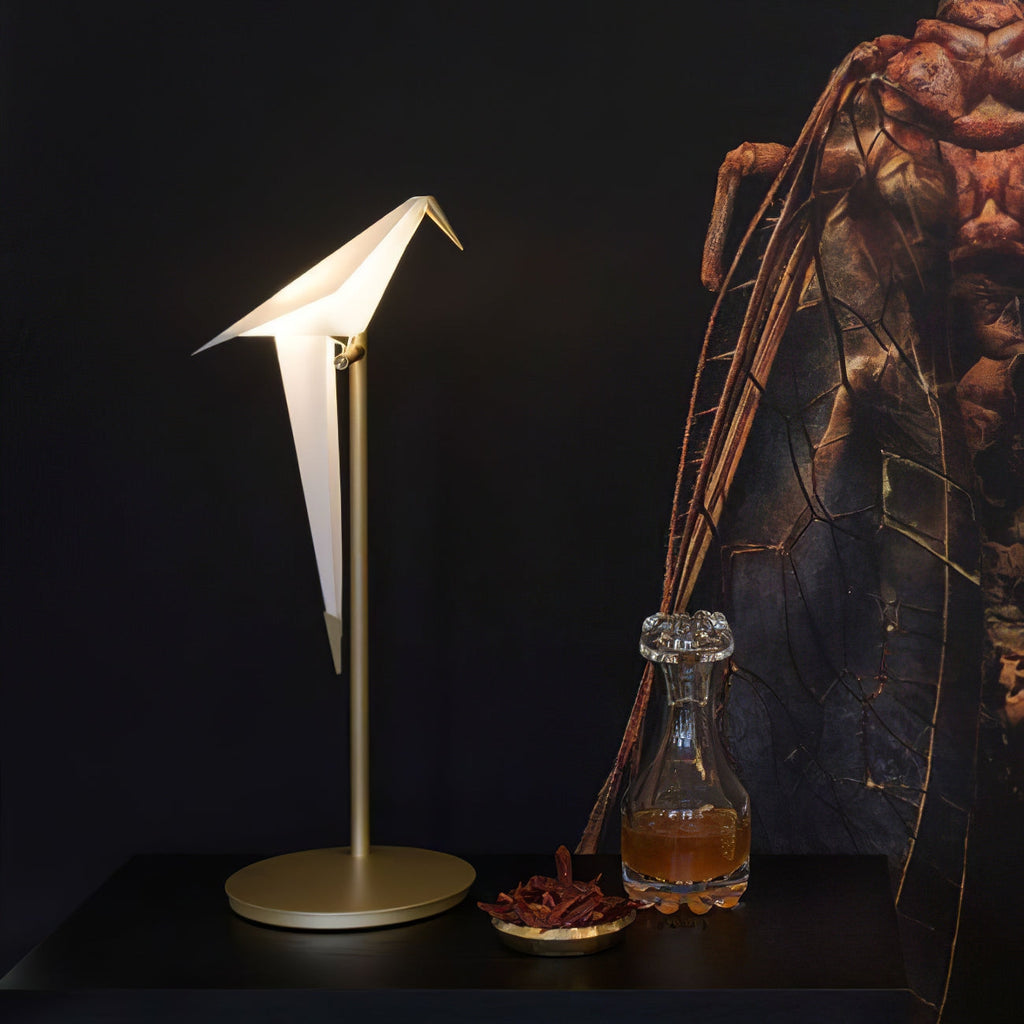 A lit Clérachic bird lamp on a gold stand and wide round base, on a dark table beside a glass decanter and a small dish.
