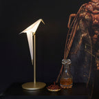 A lit Clérachic bird lamp on a gold stand and wide round base, on a dark table beside a glass decanter and a small dish.