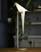 A Clérachic bird table lamp on a gold stand, on a wooden table in front of a trailing plant and grey shelves.