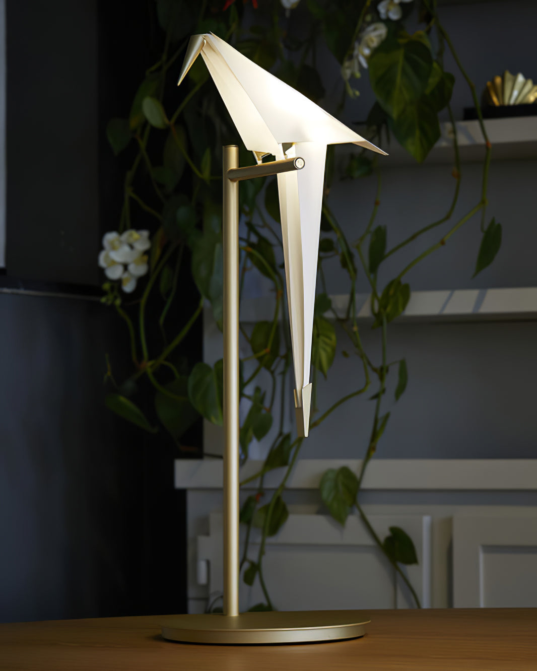 A Clérachic bird table lamp on a gold stand, on a wooden table in front of a trailing plant and grey shelves.