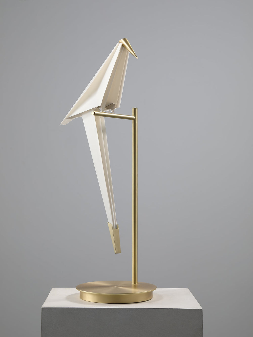 A Clérachic bird table lamp, unlit, on a gold stand and flat round base, standing on a pale concrete plinth.