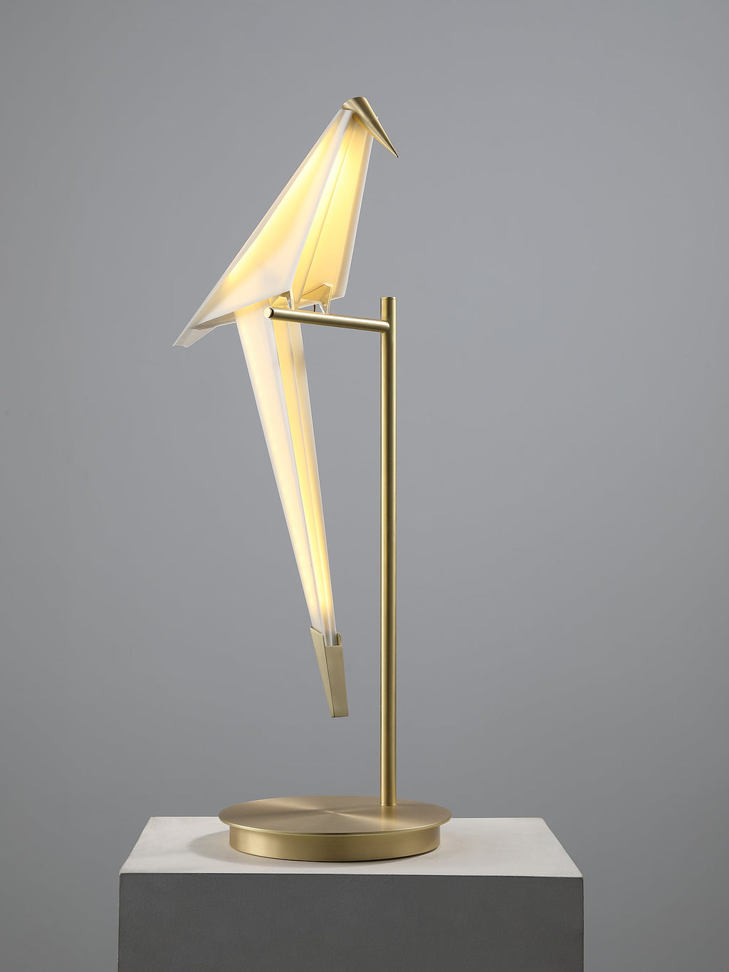 The bird of this Clérachic table lamp swings forward, lit from inside, on a gold stand on a concrete plinth.