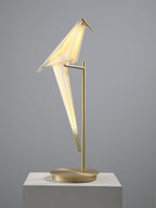 A lit Clérachic bird table lamp tilted on its gold stand, standing on a pale plinth against a grey backdrop.