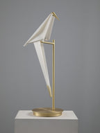 Unlit, the white folded bird of this Clérachic table lamp leans on its gold stand on a pale plinth.