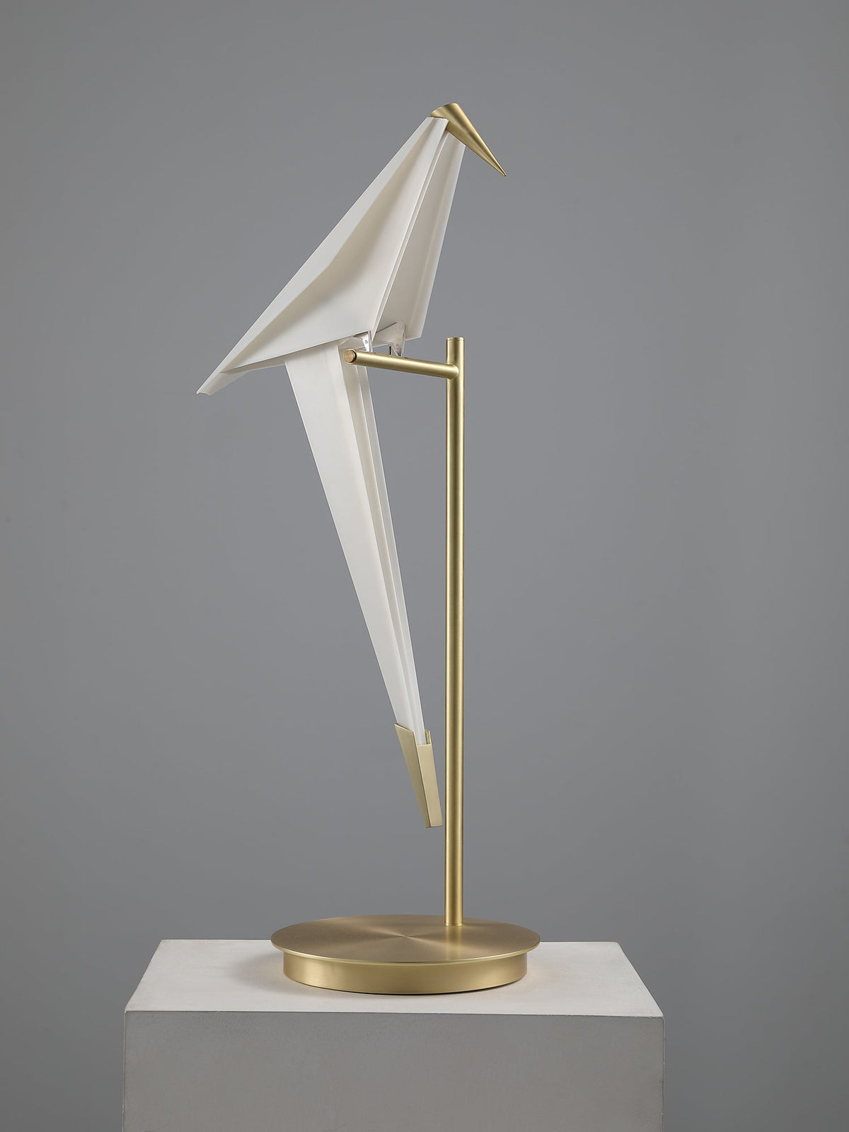Unlit, the white folded bird of this Clérachic table lamp leans on its gold stand on a pale plinth.