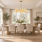 This Clérachic linear chandelier hangs in front of a tall window over a light oak dining table and upholstered chairs.