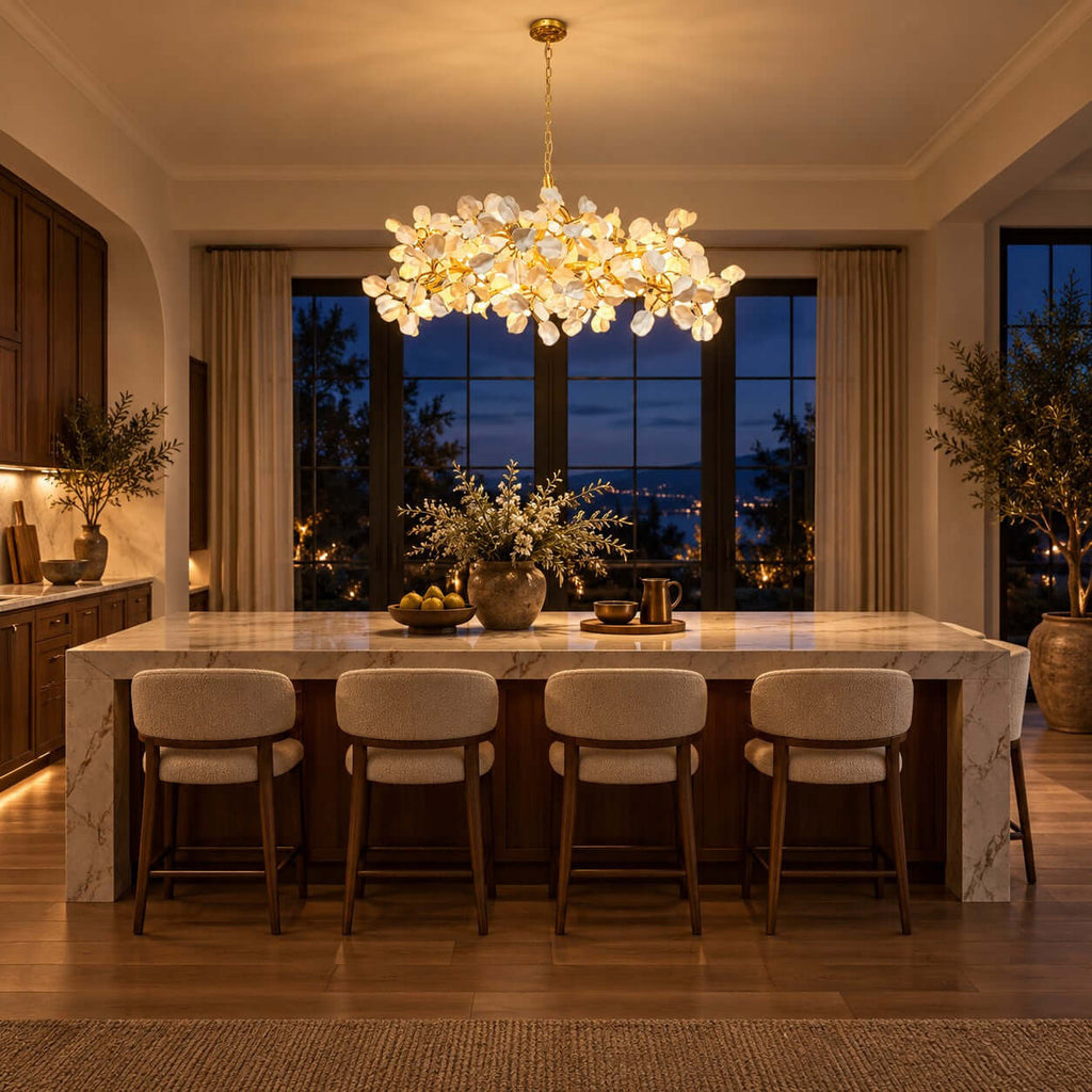 Evening kitchen with a Clérachic chandelier glowing gold above a marble island, cushioned stools and windows on a dusk sky.