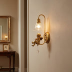 Lit Clérachic wall light with a gold cherub and clear glass bell shade, beside a gilt mirror and a dark wooden console.