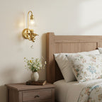 Clérachic cherub wall light with a clear shade beside a wooden bed with floral bedding, a nightstand and wildflowers.