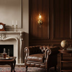 Clérachic gold cherub wall light glowing against dark wood panelling, above a leather armchair and a cream fireplace.