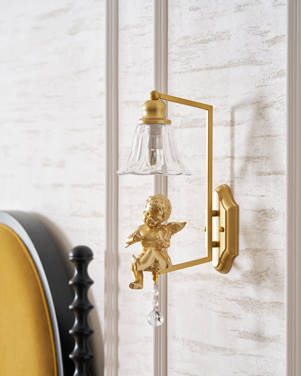 Clérachic wall light with a gold cherub, shown beside a mustard-velvet headboard against a pale panelled wall.