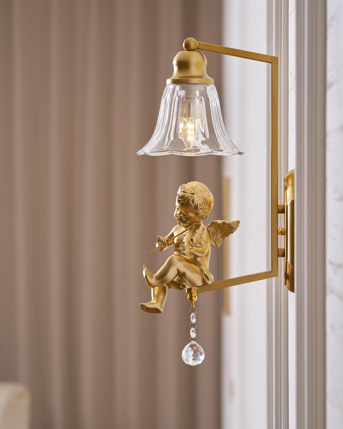 Close view of a gold cherub with a violin on a Clérachic wall light, with a crystal drop and a tan curtain behind.