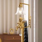 Clérachic gold cherub wall light with a square arm, with a wooden chest and a small gold frame blurred behind it.