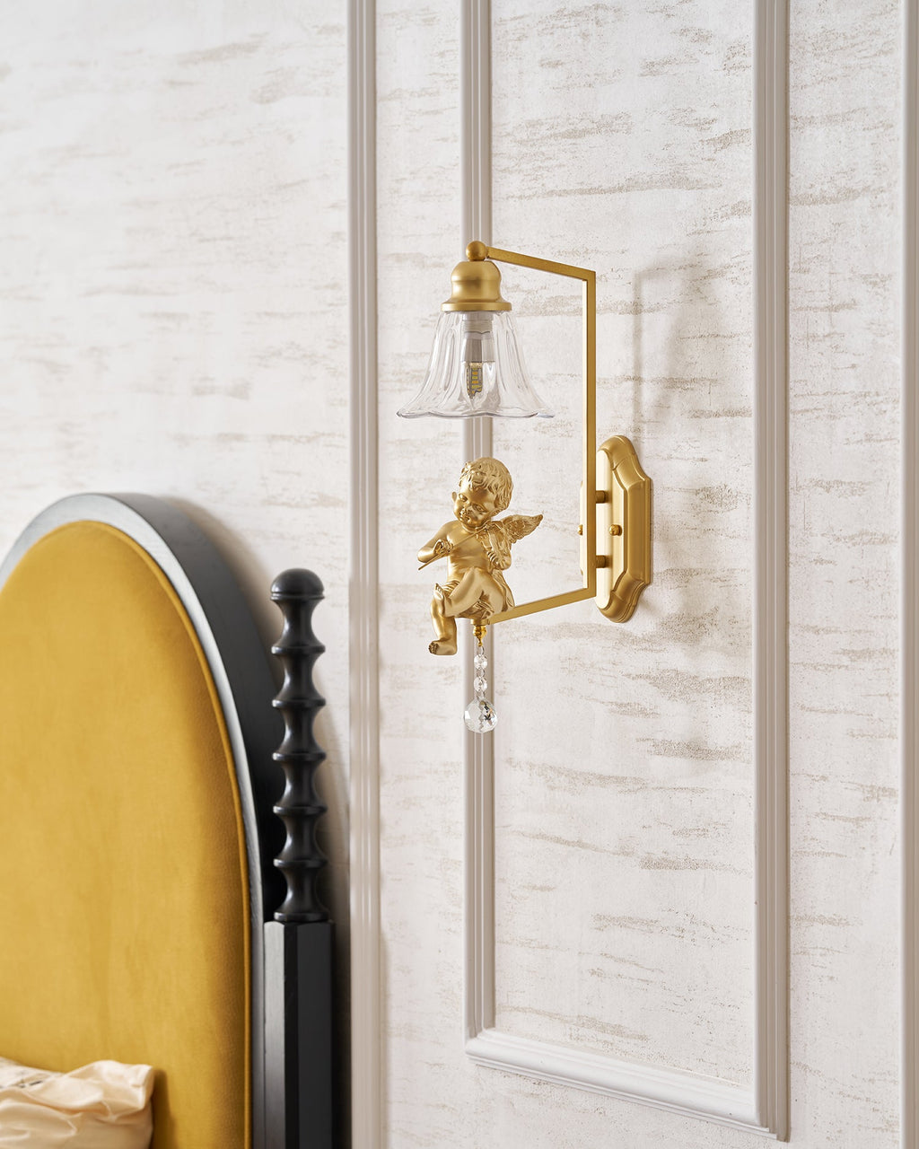 Clérachic gold cherub wall light on a white panelled wall, with the edge of a mustard headboard at the left.