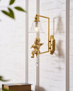 Clérachic gold cherub wall light on a white panelled wall, with a closed book and green leaves at the lower and upper edges.