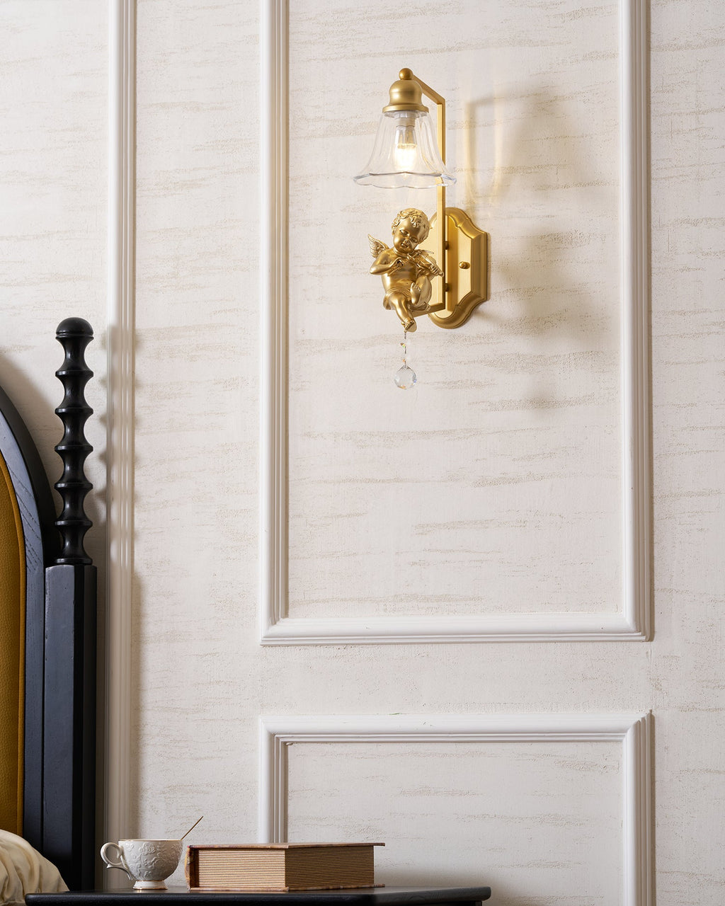 Clérachic gold cherub wall light centred on a white panel, with a black bedpost and a bedside table at the left.