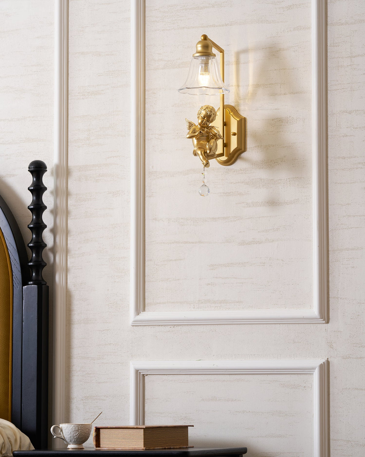 Clérachic gold cherub wall light centred on a white panel, with a black bedpost and a bedside table at the left.