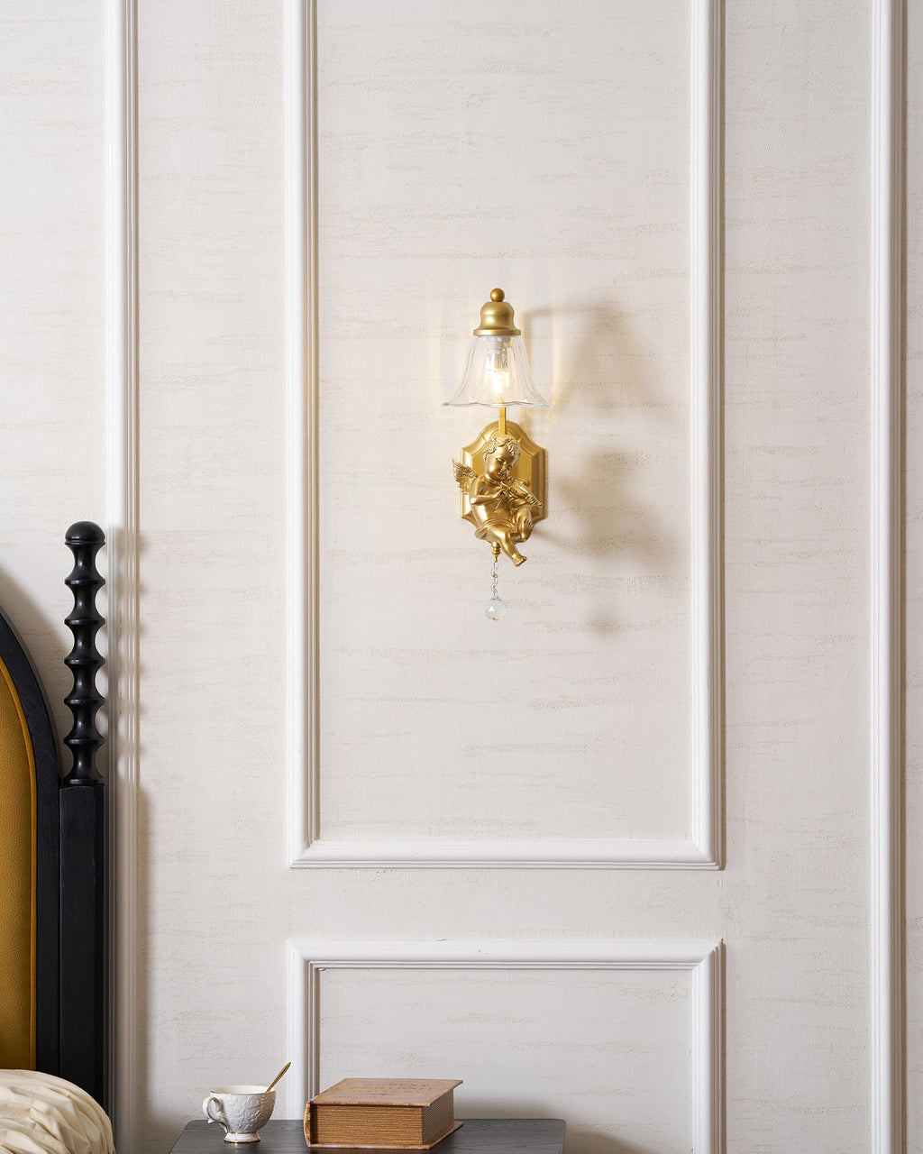 Distant view of a lit Clérachic gold cherub wall light on a white panelled wall above a bedside table.