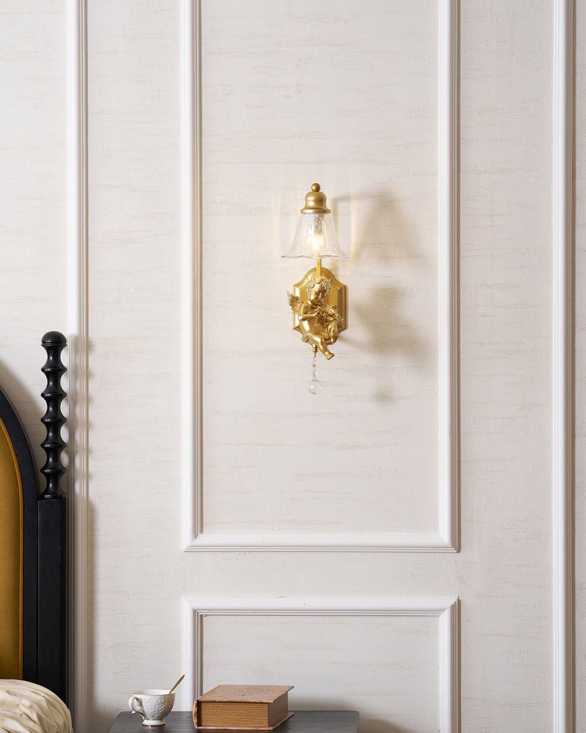 Distant view of a lit Clérachic gold cherub wall light on a white panelled wall above a bedside table.