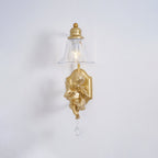 Front view of a Clérachic gold wall light, its clear bell shade above a cherub and a crystal drop on a pale grey wall.