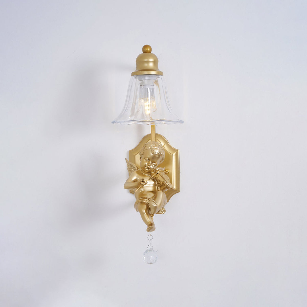 Front view of a Clérachic gold wall light, its clear bell shade above a cherub and a crystal drop on a pale grey wall.