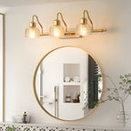 Lit three-light Clérachic wall lamp mounted above a round gold-framed bathroom mirror, with a green stem in a glass vase nearby.