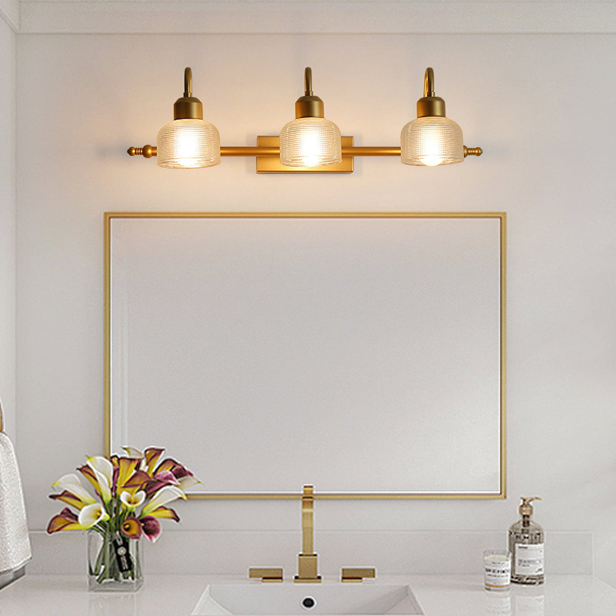 Three gold arms with warm-glowing ribbed shades sit above a rectangular gold-framed mirror and a gold tap on this Clérachic wall lamp.