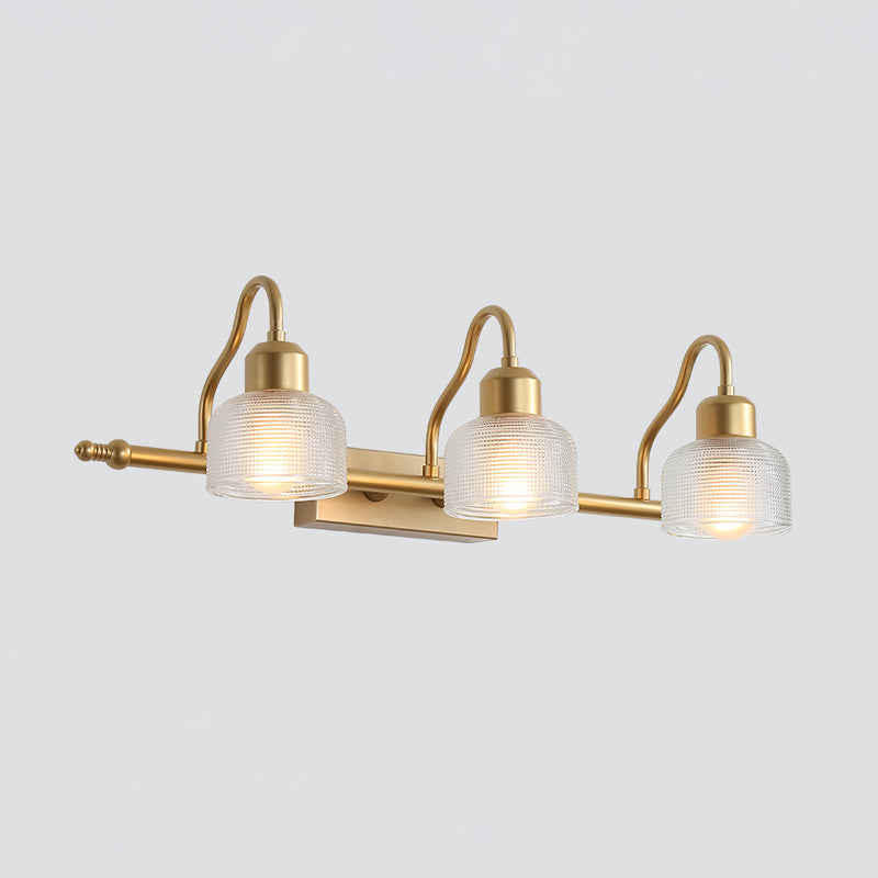 Three-light Clérachic wall lamp with clear ribbed glass shades glowing softly on a long gold bar, shown against a light grey backdrop.
