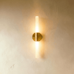 Clérachic Ena wall lamp in gold with a slim frosted glass tube, lit warmly at both ends against a plaster wall.