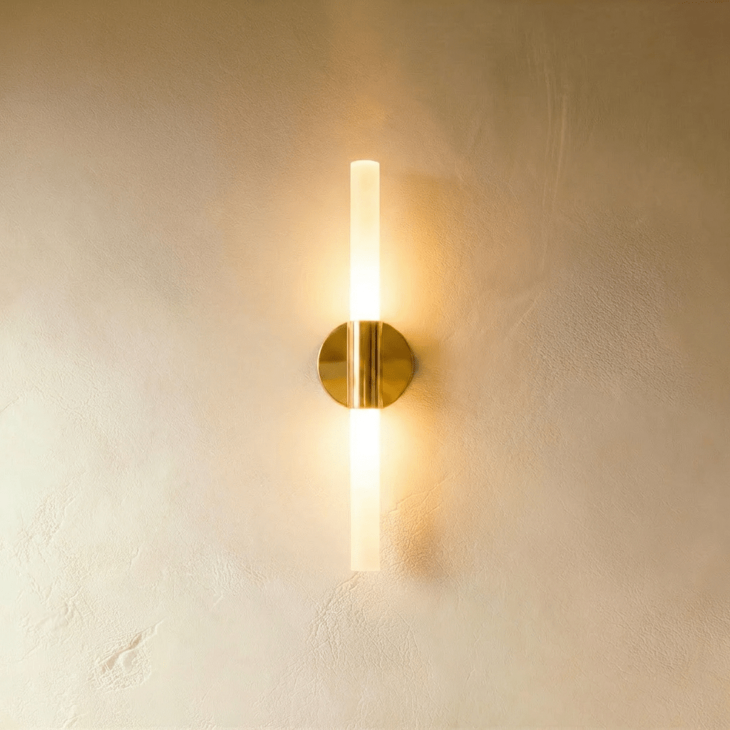Clérachic Ena wall lamp in gold with a slim frosted glass tube, lit warmly at both ends against a plaster wall.