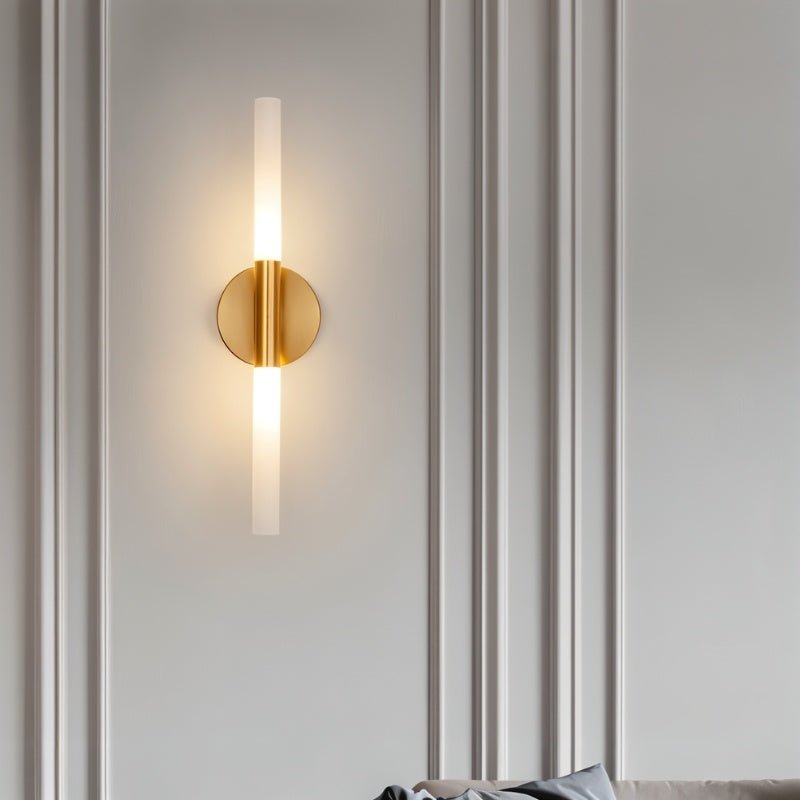 A Clérachic Ena wall lamp in gold lights a panelled white wall beside a soft grey sofa cushion.