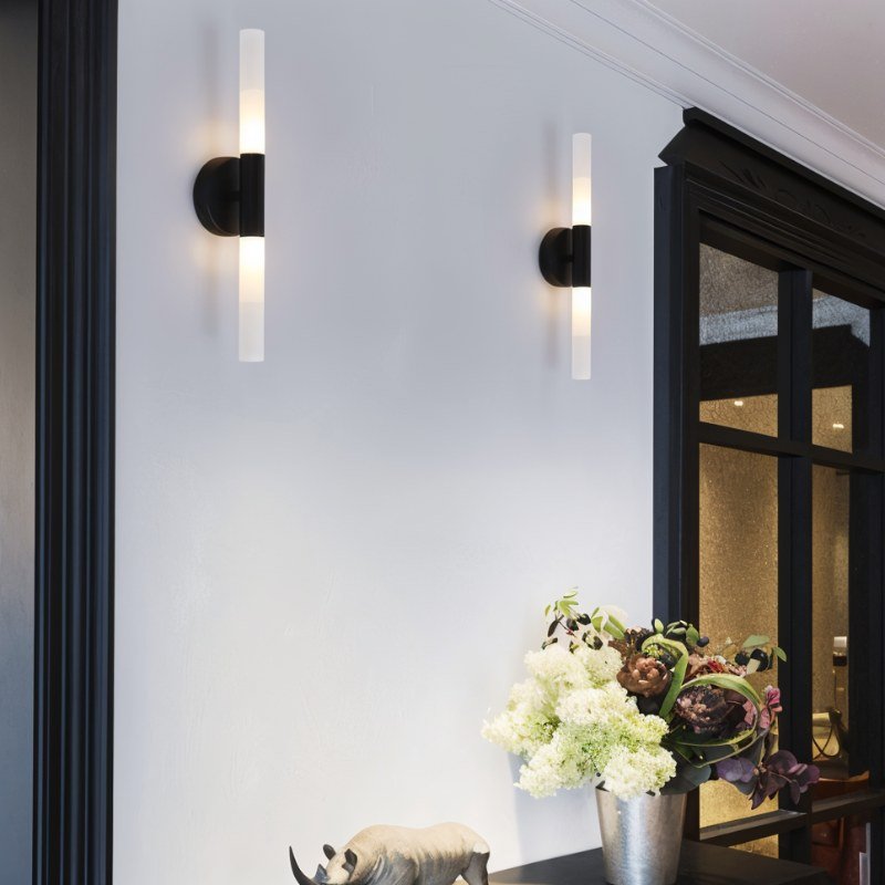 Two Clérachic Ena wall lamps in black flank a dark-framed doorway near a vase of white flowers.
