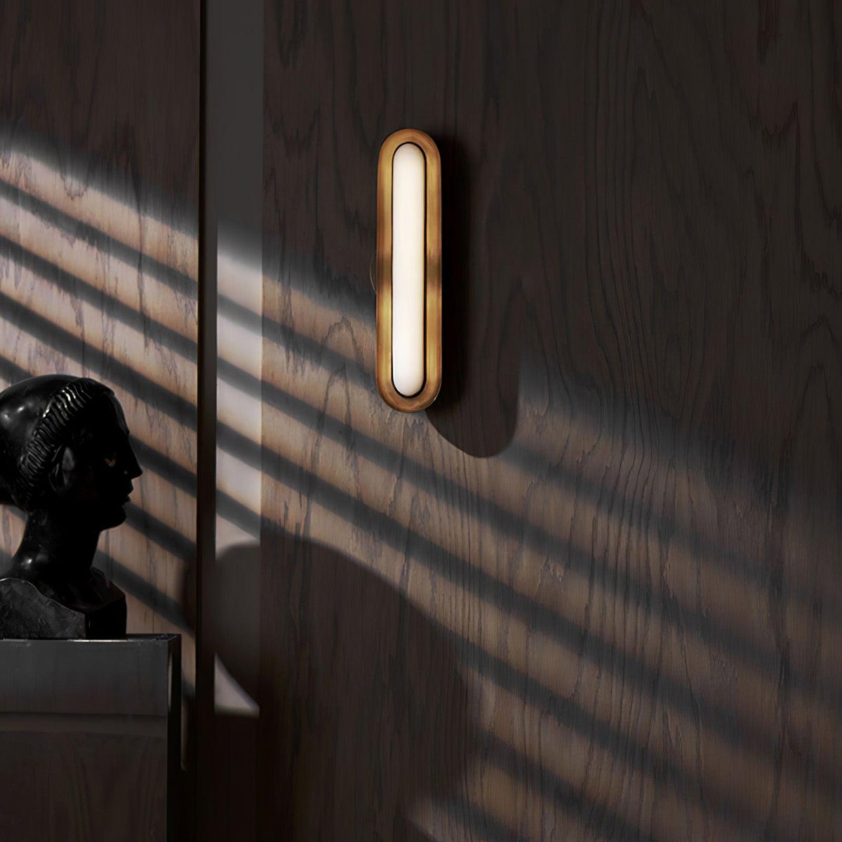 A small gold-rimmed capsule wall light with a frosted white lens on a dark oak-panelled wall, striped by sunlight, from Clérachic.