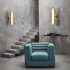 Two Clérachic capsule wall lights with gold rims sit high on a concrete wall above a floating stair and a teal armchair.