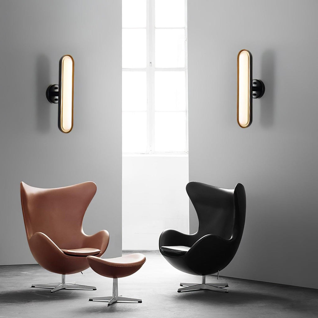 A pair of gold-rimmed capsule wall lights on a pale wall above a tan and a black egg chair, in a bright room, from Clérachic.