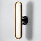 Close view of a tall frosted white capsule lens in a gold rim on a round black wall plate, against a pale wall, from Clérachic.