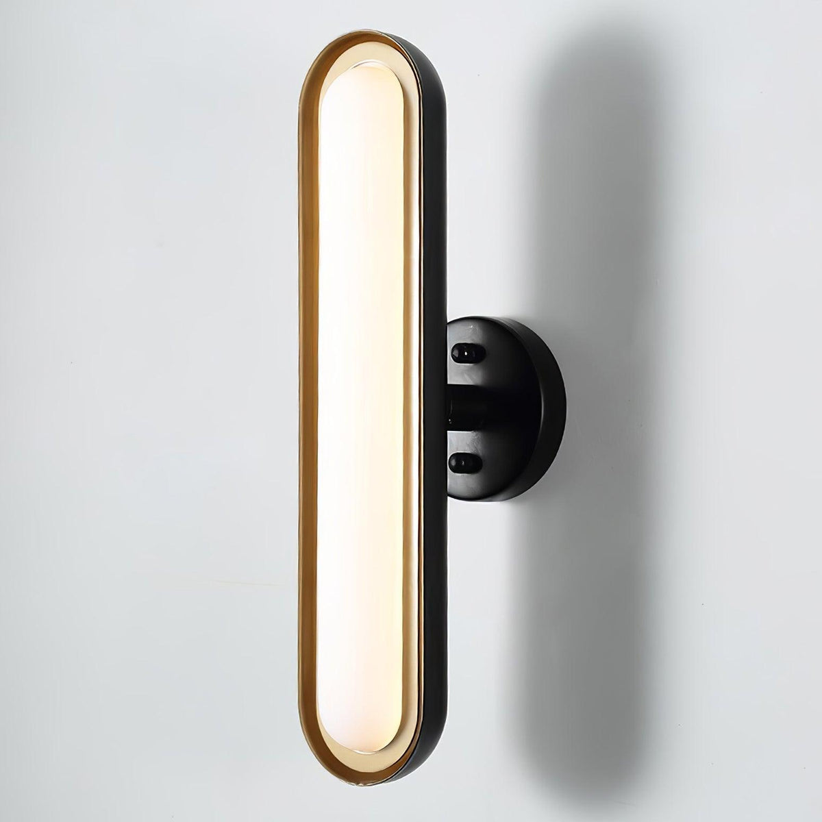 Close view of a tall frosted white capsule lens in a gold rim on a round black wall plate, against a pale wall, from Clérachic.