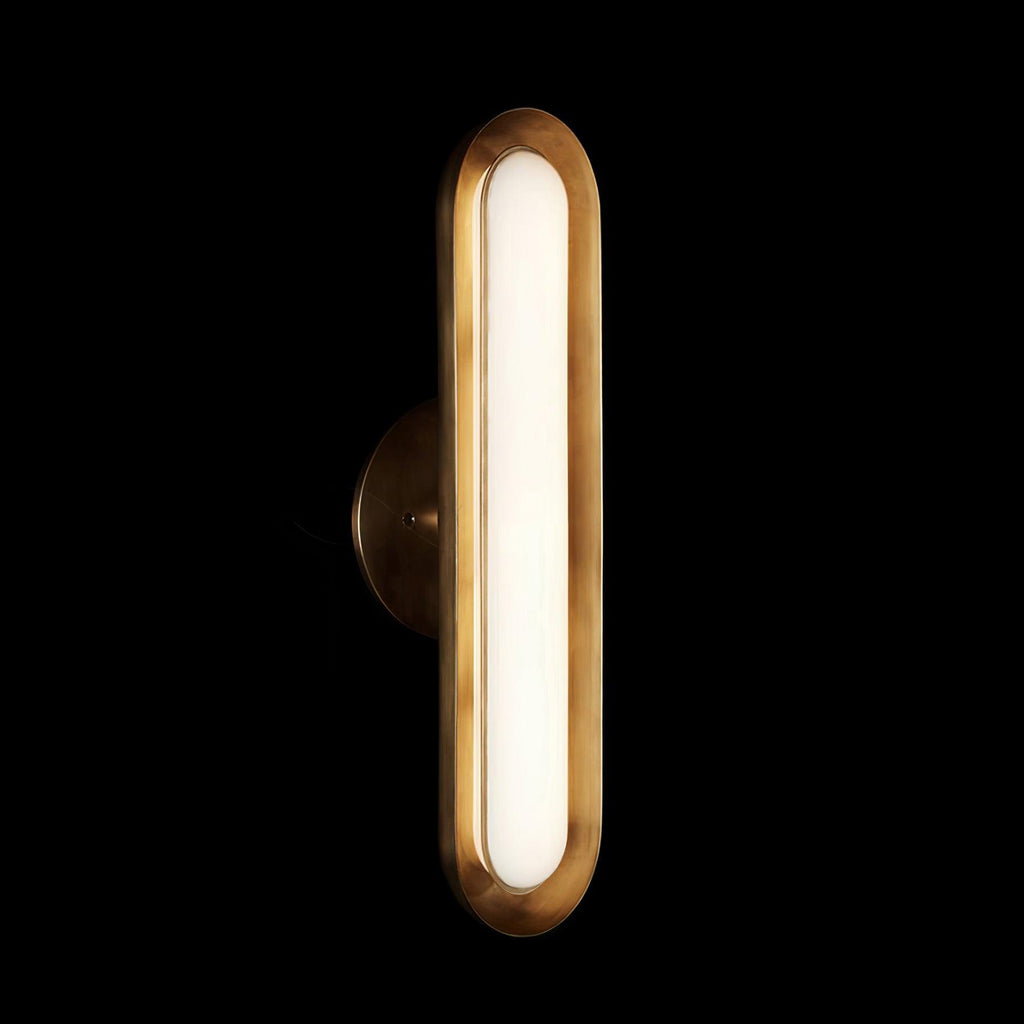 Lit against a black backdrop, this Clérachic capsule wall light shows a brushed gold rim and a glowing frosted white lens.