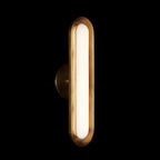 Lit against a black backdrop, this Clérachic capsule wall light shows a brushed gold rim and a glowing frosted white lens.
