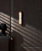 A narrow view of a Clérachic capsule wall light on dark wood panelling, with a bust and stripes of sun on the wall.