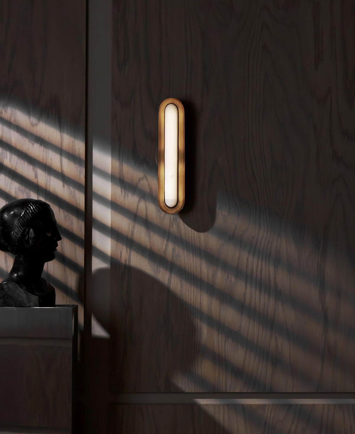A narrow view of a Clérachic capsule wall light on dark wood panelling, with a bust and stripes of sun on the wall.
