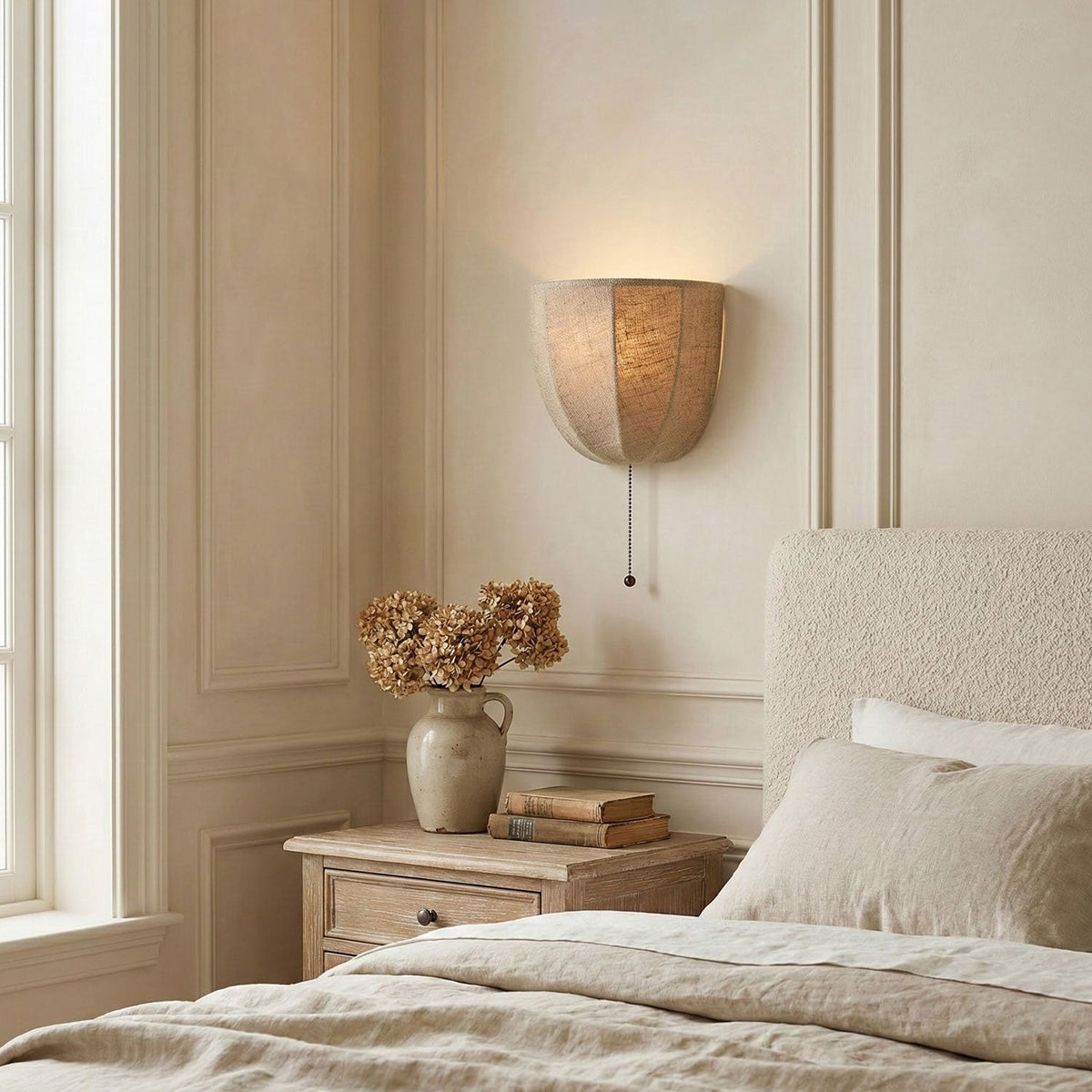 Clérachic linen wall sconce glowing softly above a bedside table, with a cream bed, dried flowers and panelled walls.