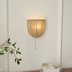 Clérachic linen wall sconce lit warm on a white wall, with a pull chain hanging below and a green sprig at the left.