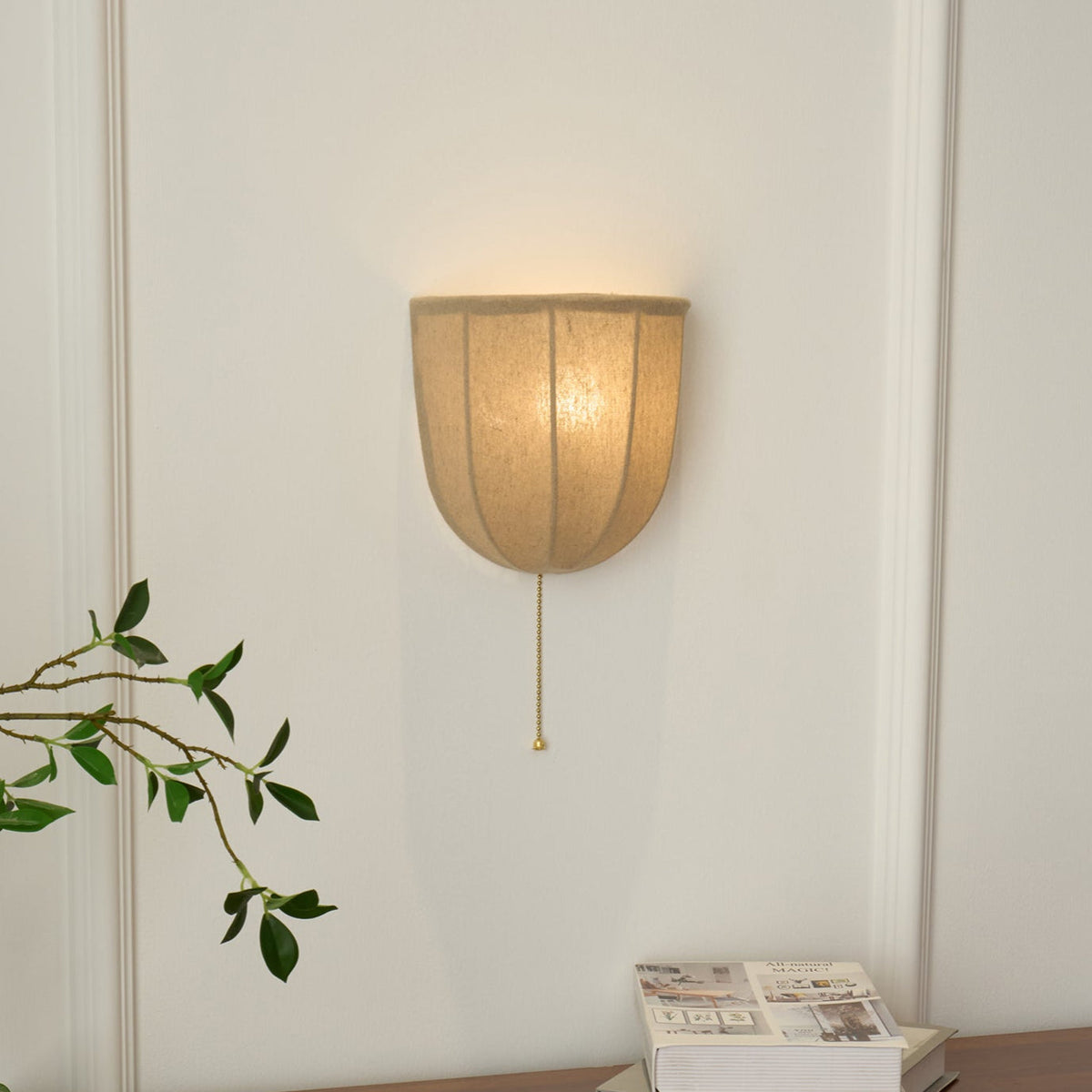 Clérachic linen wall sconce lit warm on a white wall, with a pull chain hanging below and a green sprig at the left.