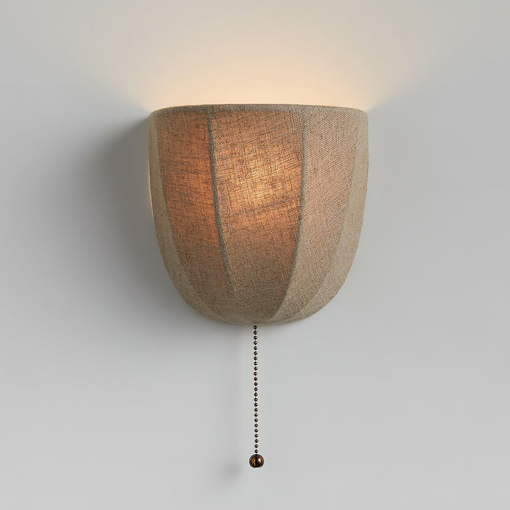 Lit Clérachic linen wall sconce on a pale grey wall, its seamed shade glowing amber with a beaded pull chain below.