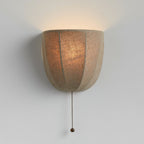 Lit Clérachic linen wall sconce on a pale grey wall, its seamed shade glowing amber with a beaded pull chain below.