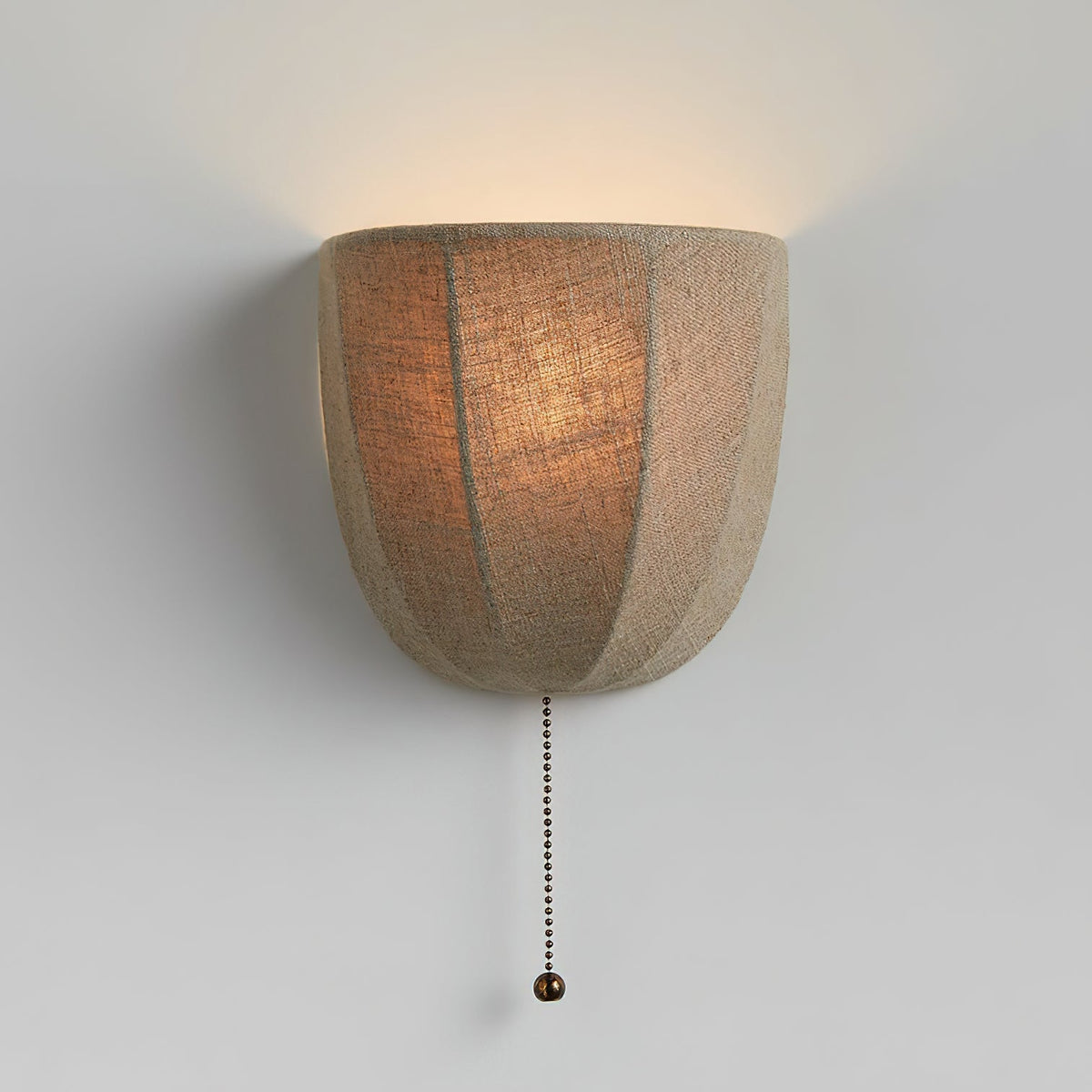 Lit Clérachic linen wall sconce on a pale grey wall, its seamed shade glowing amber with a beaded pull chain below.